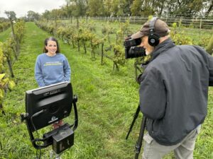A woman wearing a blue long-sleeve shirt speaks to a camera on a farm. 