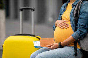 A pregnant woman sits in an airport with her flight tickets in hand. She has a yellow shirt and a denim jacket. Her suitcase is yellow. Her face is not in the picture. 