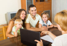 A young family sits at a wooden table, they are smiling and listening while a well-dressed person across from them gestures to a laptop screen. It is a classroom setting.