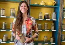 Local Shop Explores Natural Remedies for Family Health A woman holds a jar of herbs. She is smiling. She stands in front of a shelf with similar jars containing a variety of herbs.
