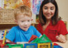 A teacher helps a child play with magnetic block pieces. The teacher is wearing a red polo and has shoulder-length brown hair. The child has light colored hair and a blue T-shirt.