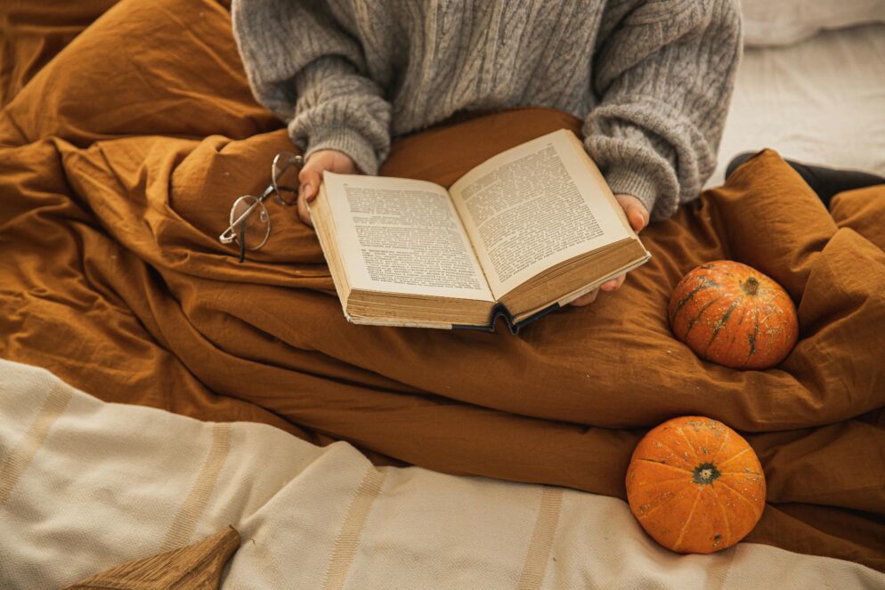 A women sits wrapped in cozy blankets holding a book with mini pumpkins placed around her. You cannot see her face. This is to show the theme of books about Halloween.
