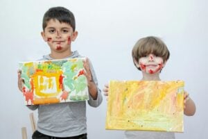 Two children holding up paintings, their faces are covered in red paint