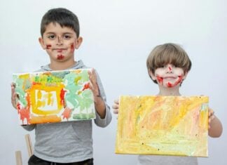 Two children holding up paintings, their faces are covered in red paint